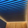 Nowoczesna pergola z oświetleniem LED do pergoli lamelowej, poziomymi metalowymi listwami i ciepłym oświetleniem taśmowym wzdłuż krawędzi, przymocowana do drewnianej ściany i sfotografowana o zmierzchu z błękitnym niebem prześwitującym przez szczeliny.