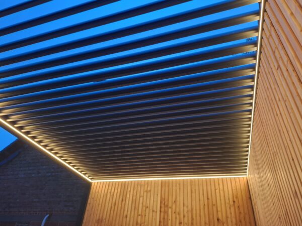 Nowoczesna pergola z oświetleniem LED do pergoli lamelowej, poziomymi metalowymi listwami i ciepłym oświetleniem taśmowym wzdłuż krawędzi, przymocowana do drewnianej ściany i sfotografowana o zmierzchu z błękitnym niebem prześwitującym przez szczeliny.