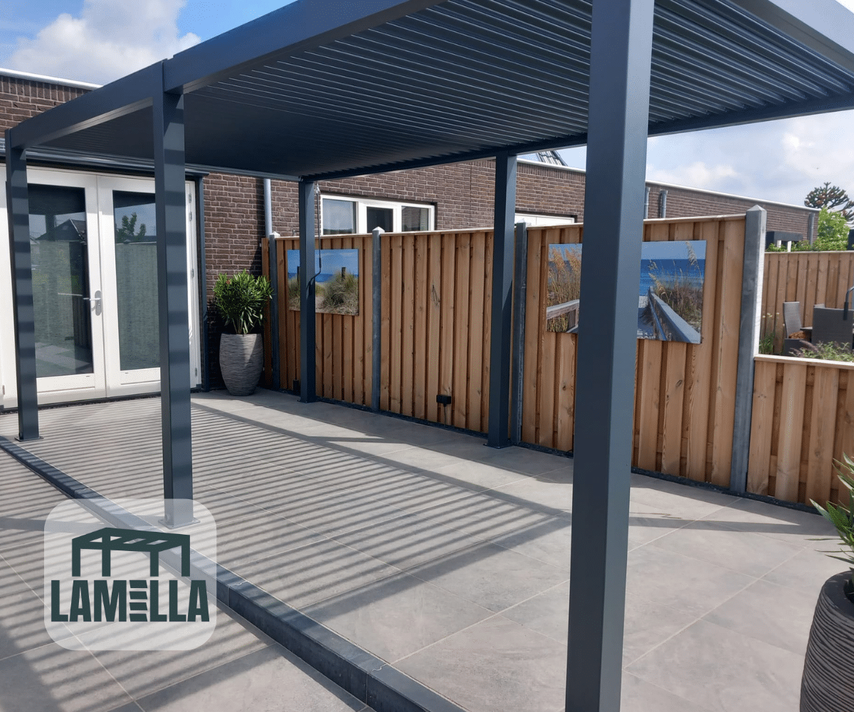 Nowoczesne patio posiada Elektryczne zadaszenie lamelowe jako elegancką szarą pergolę, z dużymi płytkami i drewnianym płotem prezentującym dzieła sztuki na świeżym powietrzu. Szklane drzwi i rośliny doniczkowe dodają stylu. Logo Lamella znajduje się w lewym dolnym rogu.