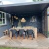 Nowoczesne zadaszone patio z drewnianym stołem, sześcioma czarnymi krzesłami, wiszącymi dekoracjami, lampkami sznurkowymi i oprawioną sztuką. Szewronowe drewniane podłogi i bujne rośliny podkreślają przestrzeń pod stylową przesuwną ścianą lamelową lub stałym panelem lamelowym.