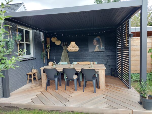 Nowoczesne zadaszone patio z drewnianym stołem, sześcioma czarnymi krzesłami, wiszącymi dekoracjami, lampkami sznurkowymi i oprawioną sztuką. Szewronowe drewniane podłogi i bujne rośliny podkreślają przestrzeń pod stylową przesuwną ścianą lamelową lub stałym panelem lamelowym.