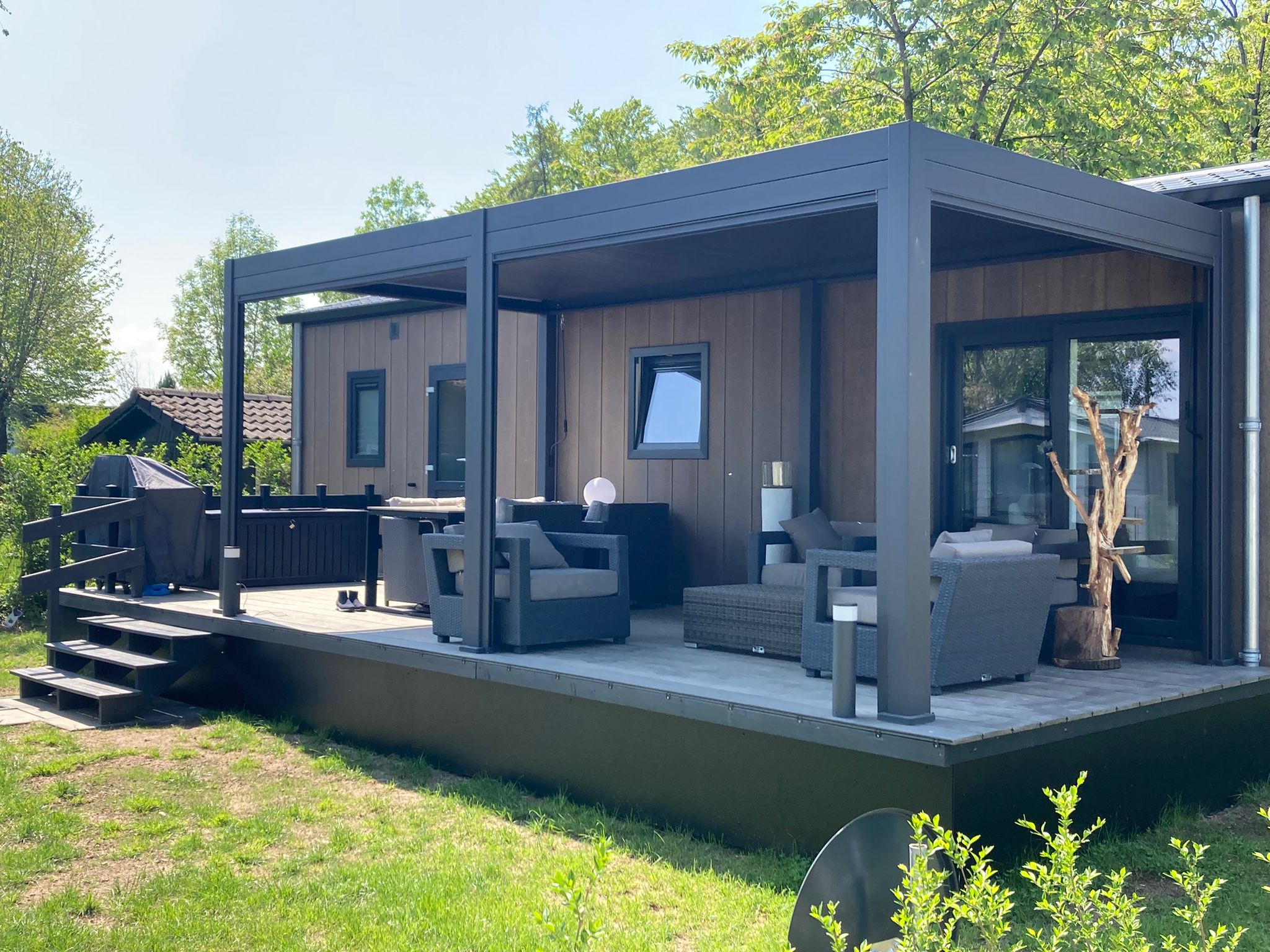 Nowoczesne patio na świeżym powietrzu z szarymi meblami, przykryte elegancką, czarną pergolą z dachem lamelowym elektrycznym, przylegającym do domu z dużymi oknami. Otoczone zielenią i drzewami, z drewnianymi schodami prowadzącymi na taras.