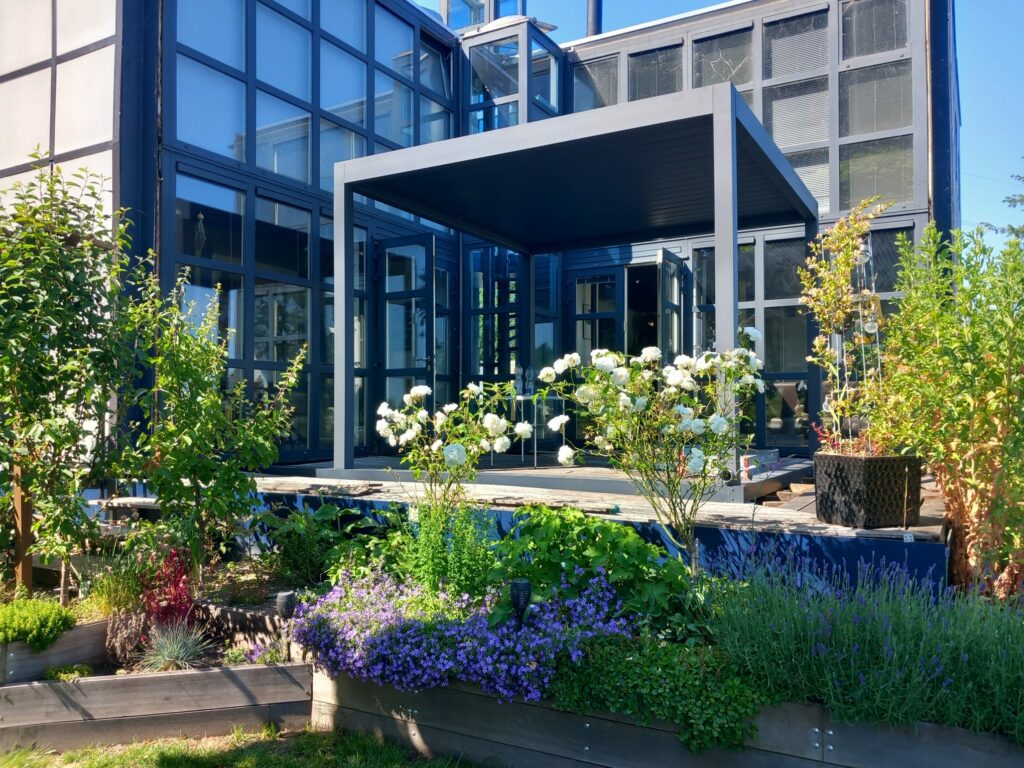 Nowoczesny szklany budynek z pergolą z dachem lamelowym elektrycznym na drewnianym pokładzie, otoczony bujnymi rabatami ogrodowymi wypełnionymi kwitnącymi roślinami i zielenią w słoneczny dzień.