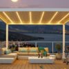 Nowoczesne patio na dachu o zmierzchu oferuje Oświetlenie LED do pergoli lamelowej na wymiar z ciepłym światłem, przytulną sofę segmentową z kolorowymi poduszkami, stół z napojami, rośliny doniczkowe i wysokie krzesło z widokiem na malownicze jezioro i góry.