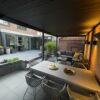 Nowoczesne zadaszone patio z zadaszeniem tarasu, wyposażone w stół jadalny, krzesła i sofę zewnętrzną. Wychodzi na przestronny dziedziniec z roślinami doniczkowymi i dużymi oknami na ceglanym domu. Ciepłe oświetlenie tworzy przytulną atmosferę.