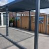 Nowoczesne patio z dużymi szarymi płytkami, eleganckie zadaszenie tarasu z dachem lamelowym elektrycznym rzucającym pasiaste cienie, drewniane ogrodzenie ze sztuką na zewnątrz i rośliny doniczkowe obok szklanych drzwi prowadzących do ceglanego budynku.