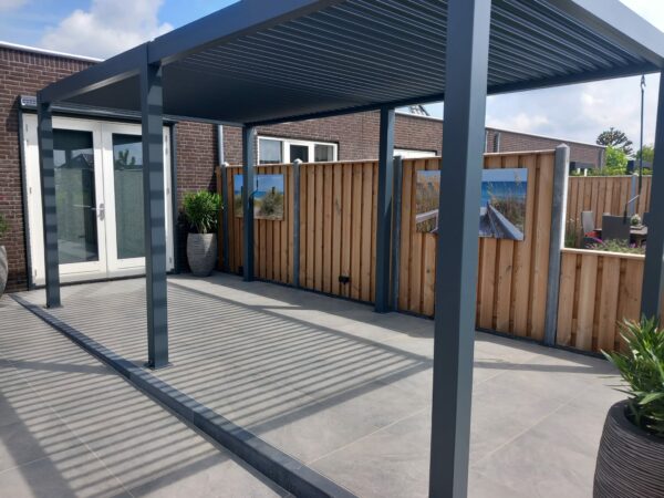 Nowoczesne patio z dużymi szarymi płytkami, eleganckie zadaszenie tarasu z dachem lamelowym elektrycznym rzucającym pasiaste cienie, drewniane ogrodzenie ze sztuką na zewnątrz i rośliny doniczkowe obok szklanych drzwi prowadzących do ceglanego budynku.