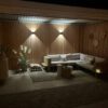 Przytulne patio na świeżym powietrzu nocą obejmuje drewnianą narożną sofę z poduszkami pod elegancką pergolą, mały stolik kawowy, rośliny doniczkowe, oświetlenie ścienne i dekoracyjne akcenty ścienne na tle drewnianych paneli ściennych.