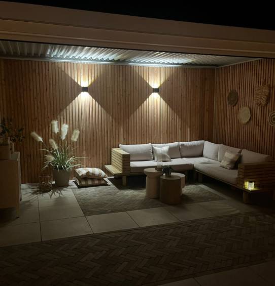 Przytulne patio na świeżym powietrzu nocą obejmuje drewnianą narożną sofę z poduszkami pod elegancką pergolą, mały stolik kawowy, rośliny doniczkowe, oświetlenie ścienne i dekoracyjne akcenty ścienne na tle drewnianych paneli ściennych.