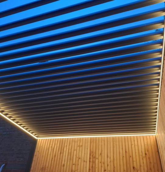 Nowoczesna drewniana pergola z poziomymi listwami i wbudowanym oświetleniem LED świeci ciepło o zmierzchu, podkreślając fakturę drewna i kontrastując z błękitnym wieczornym niebem nad nią - stylowe zadaszenie tarasu dla każdego domu.