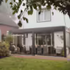 Nowoczesne, przeszklone patio z wiklinowymi krzesłami i stołem jadalnym rozciąga się z tyłu białego domu, wyposażonego w elegancki dach lamelowy elektryczny i otoczonego zielonym ogrodem i drzewami.