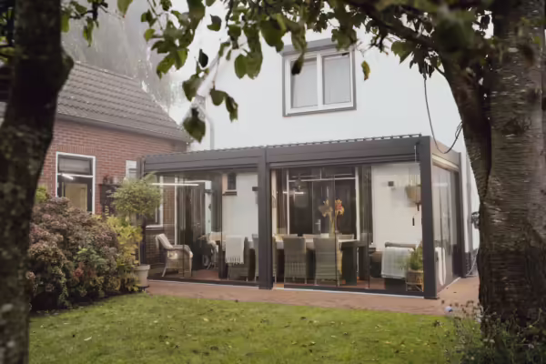 Nowoczesne, przeszklone patio z wiklinowymi krzesłami i stołem jadalnym rozciąga się z tyłu białego domu, wyposażonego w elegancki dach lamelowy elektryczny i otoczonego zielonym ogrodem i drzewami.