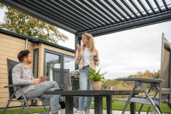 Mężczyzna siedzi na krześle trzymając tablet, podczas gdy kobieta stoi obok rozmawiając i trzymając filiżankę na nowoczesnym patio z pergolą, otoczonym roślinami i wygodnymi siedzeniami.