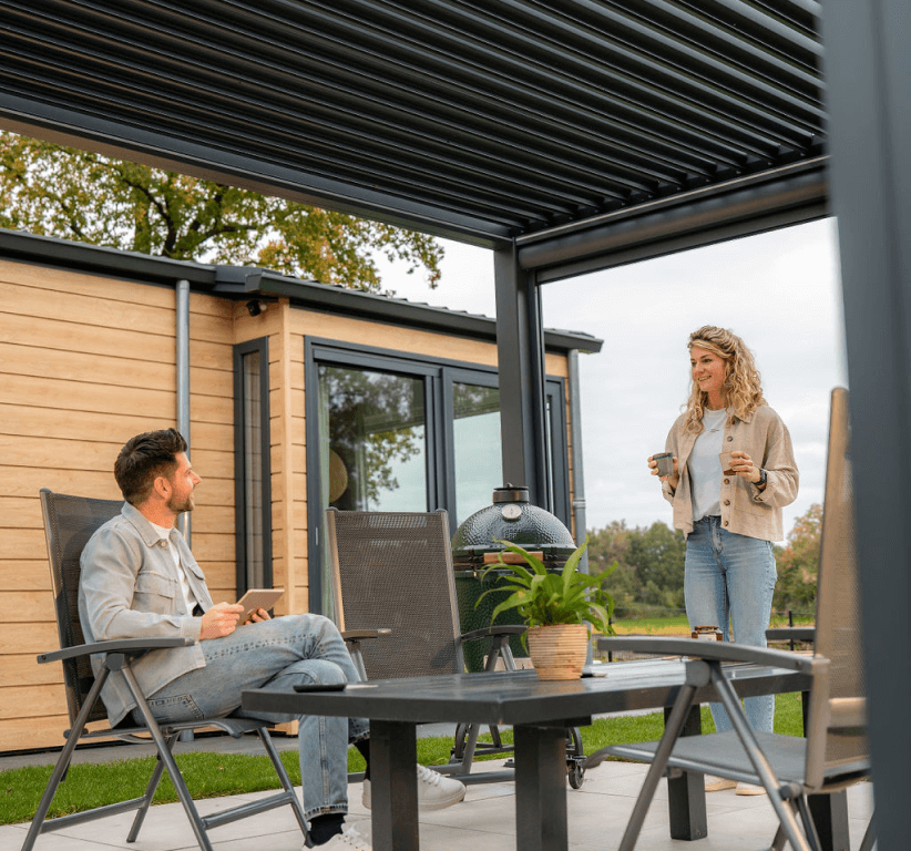 Mężczyzna siedzi przy stole na patio, podczas gdy kobieta stoi trzymając dwa kubki, oboje ciesząc się rozmową pod elegancką pergolą z elektrycznym dachem lamelowym na podwórku z drewnianym domem i zielonym trawnikiem.