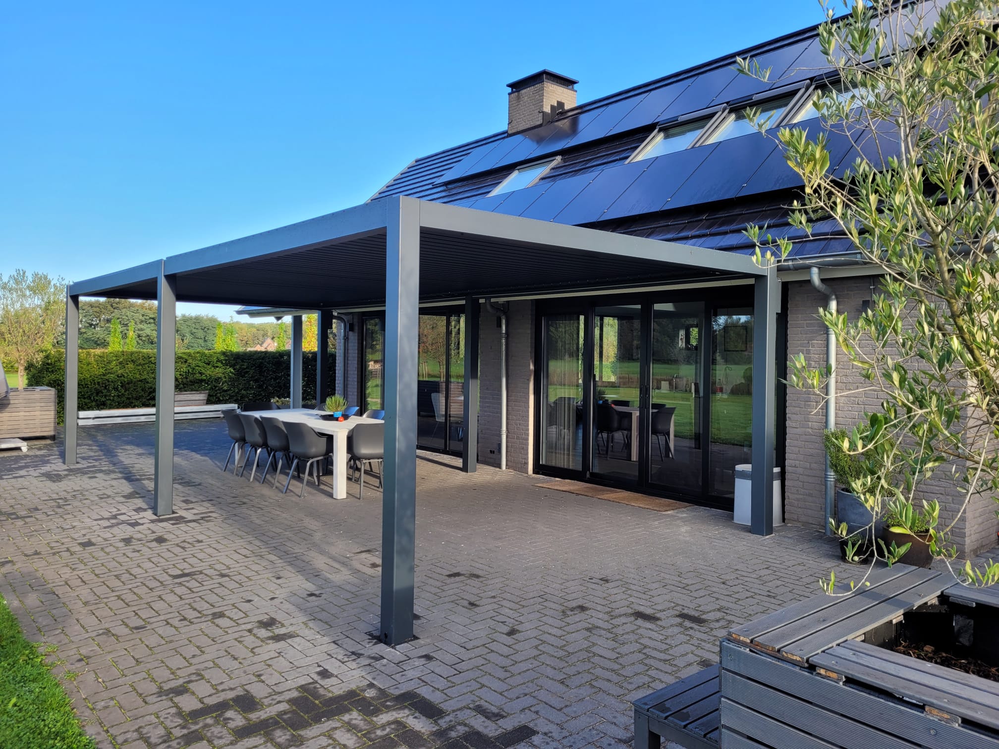 Nowoczesne patio z metalową pergolą lub dachem lamelowym obejmuje stół i krzesła na świeżym powietrzu obok domu z dużymi szklanymi drzwiami i panelami słonecznymi, otoczone brukowanym tarasem i zielenią.