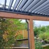 Drewniana pergola z dachem lamelowym elektrycznym stoi obok stawu otoczonego bujną zielenią, roślinami i drewnianym płotem w pochmurny dzień.