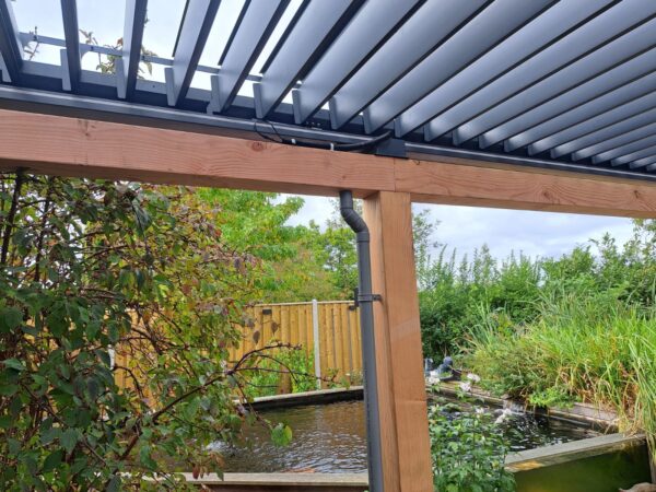 Drewniana pergola z dachem lamelowym elektrycznym stoi obok stawu otoczonego bujną zielenią, roślinami i drewnianym płotem w pochmurny dzień.