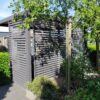 Nowoczesna pergola ogrodowa z poziomymi szarymi listwami stoi wśród bujnej zieleni i drzew, rzucając pasiaste cienie na ziemię w jasnym świetle słonecznym.