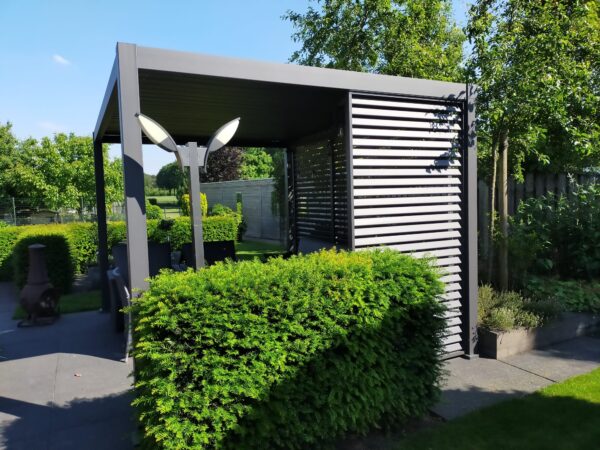 Nowoczesna altana zewnętrzna z dachem lamelowym elektrycznym stoi na patio otoczonym starannie przyciętymi żywopłotami, drzewami i zielenią w słoneczny dzień. W pobliżu widoczny jest słup latarni.