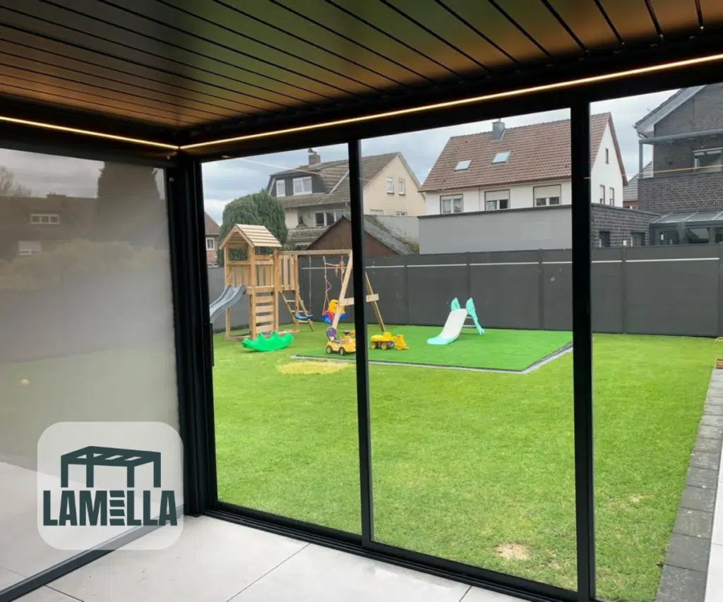 Ciesz się widokiem z zadaszonego patio ze szklanymi przesuwanymi drzwiami i pergolą Lamellendach manuell, z widokiem na trawiaste podwórko z konstrukcją do zabawy, huśtawkami, zjeżdżalnią, domkami i szarym ogrodzeniem. Logo Lamella w lewym dolnym rogu.