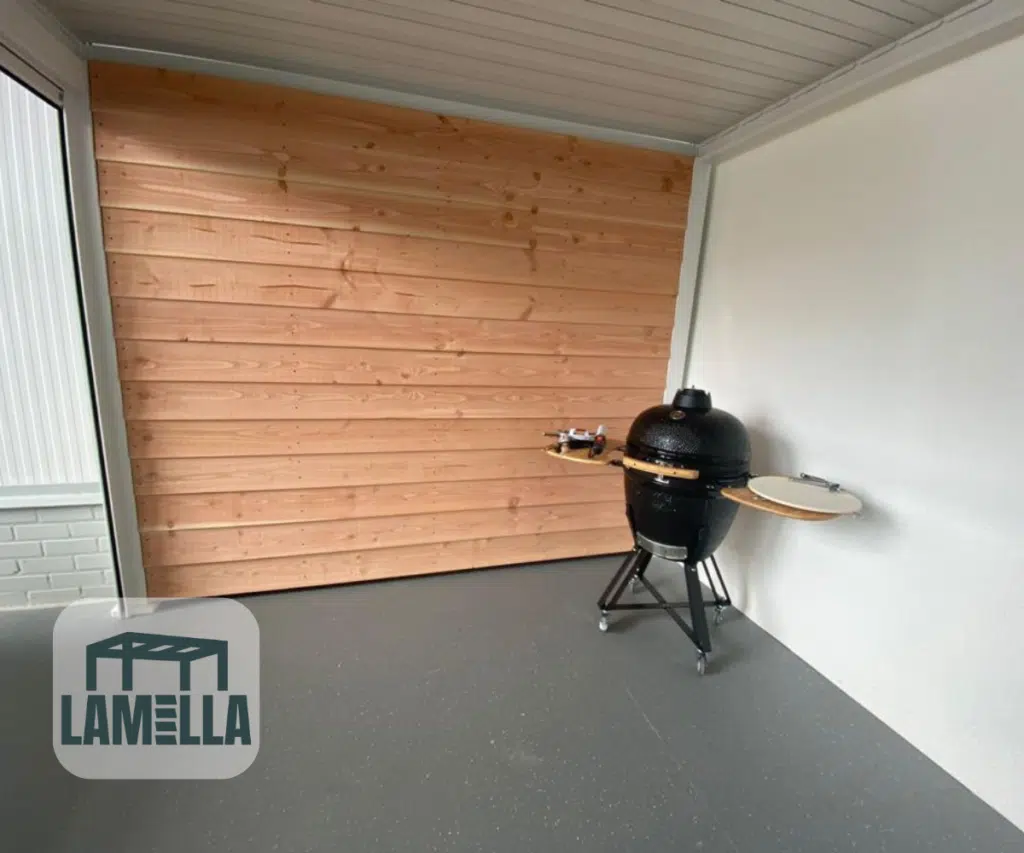 Model Lamellendach manuell (kopia) jest zainstalowany na zadaszonym patio z białymi i drewnianymi ścianami, osłaniając czarny grill zewnętrzny ze stolikami bocznymi na szarej podłodze. Logo Lamella znajduje się w lewym dolnym rogu.