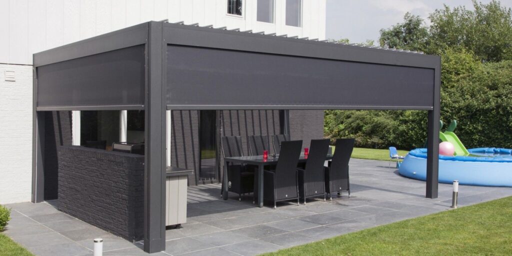 Nowoczesne patio na świeżym powietrzu z eleganckim, prostokątnym dachem lamelowym elektrycznym obejmującym stół jadalny i krzesła na podłodze wyłożonej kafelkami; zadaszenie tarasu otacza przestrzeń, z trawnikiem, nadmuchiwanym basenem i zielenią w tle.