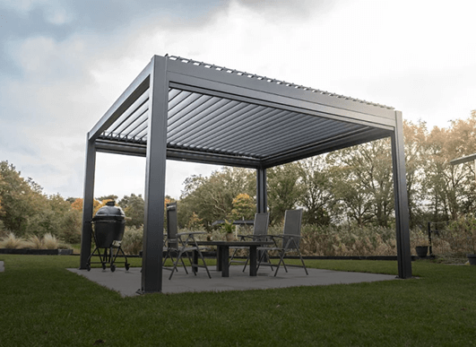 Nowoczesna pergola z dachem lamelowym elektrycznym przykrywa zestaw patio ze stołem, czterema krzesłami i grillem, a wszystko to znajduje się na trawiastym trawniku z drzewami w tle.