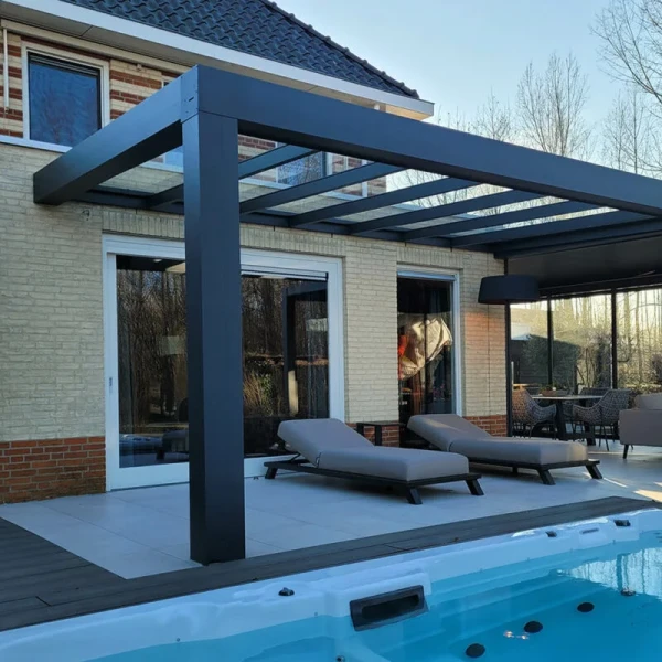 Nowoczesne patio ze szklaną pergolą Zadaszenie tarasowe Cube, dwoma szarymi fotelami i przesuwanymi drzwiami do domu. Z przodu znajduje się czysty basen, a drzewa i meble ogrodowe podkreślają tę elegancką przestrzeń.