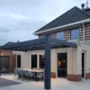 Nowoczesne patio z zestawem jadalnym znajduje się pod eleganckim, ciemnym Zadaszeniem tarasowym Cube przymocowanym do ceglanego domu. Na pergoli wiszą światła sznurkowe, a duże szklane drzwi prowadzą do środka. Niebo jest zachmurzone.