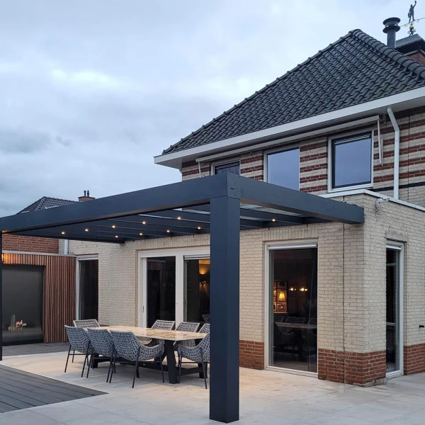 Nowoczesne patio z zestawem jadalnym znajduje się pod eleganckim, ciemnym Zadaszeniem tarasowym Cube przymocowanym do ceglanego domu. Na pergoli wiszą światła sznurkowe, a duże szklane drzwi prowadzą do środka. Niebo jest zachmurzone.