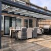 Nowoczesne patio z Zadaszeniem tarasowym ze szkła, stołem dla sześciu osób i grillem gazowym na kamiennych płytkach obok ceglanego domu z przesuwanymi szklanymi drzwiami i eleganckim dachem lamelowym elektrycznym.