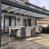 Na nowoczesnym patio znajduje się Zadaszenie tarasowe ze szkła pergola, stół jadalny na świeżym powietrzu z sześcioma wyściełanymi krzesłami i czarny grill na podłodze wyłożonej kafelkami obok ceglanego domu z dużymi przesuwanymi szklanymi drzwiami.