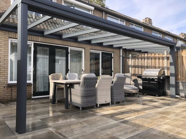 Na nowoczesnym patio znajduje się Zadaszenie tarasowe ze szkła pergola, stół jadalny na świeżym powietrzu z sześcioma wyściełanymi krzesłami i czarny grill na podłodze wyłożonej kafelkami obok ceglanego domu z dużymi przesuwanymi szklanymi drzwiami.