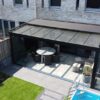 Nowoczesne podwórko z Zadaszeniem tarasowym ze szkła przykrywającym patio, z okrągłym stołem i krzesłami pod szklanym baldachimem, obok basenu, zielonego trawnika, roślin doniczkowych i ceglanego domu z dużymi oknami.
