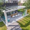 Nowoczesne przeszklone patio z Zadaszeniem tarasowym Cube wyposażone jest w długi drewniany stół jadalny dla dwóch osób, ustawiony wśród zieleni i drzew, i jest połączony z nowoczesnym domem z dużymi oknami.