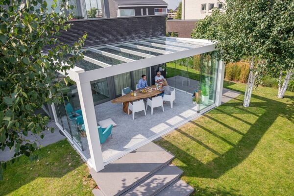Nowoczesne przeszklone patio z Zadaszeniem tarasowym Cube wyposażone jest w długi drewniany stół jadalny dla dwóch osób, ustawiony wśród zieleni i drzew, i jest połączony z nowoczesnym domem z dużymi oknami.