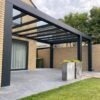 Nowoczesne patio z pergolą Zadaszenie tarasowe Cube z dachem lamelowym elektrycznym, dużymi szarymi płytkami, wysokimi ceglanymi ścianami, dwiema kwadratowymi donicami, trawnikiem i widokiem na drzewa pod zachmurzonym niebem.