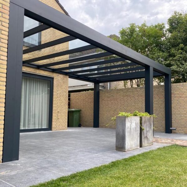 Nowoczesne patio z pergolą Zadaszenie tarasowe Cube z dachem lamelowym elektrycznym, dużymi szarymi płytkami, wysokimi ceglanymi ścianami, dwiema kwadratowymi donicami, trawnikiem i widokiem na drzewa pod zachmurzonym niebem.