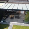 Nowoczesne patio na świeżym powietrzu z Zadaszeniem tarasowym ze szkła, szklanymi ścianami, narożną sofą, stolikiem kawowym, dekoracyjną rzeźbą białego niedźwiedzia, bujną zielenią i palmą pod elegancką pergolą.