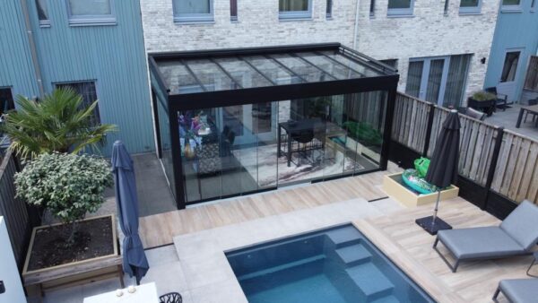 Nowoczesne podwórko z przeszklonym patio z eleganckim Zadaszenie tarasowe ze szkła, stół jadalny wewnątrz, basen, leżaki, rośliny doniczkowe i drewniane ogrodzenia obok współczesnych domów.