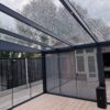 Zadaszenie tarasowe ze szkła ma nowoczesny szklany dach z kroplami deszczu, czarne metalowe ramy i przesuwane drzwi otwierające się na patio z miejscami do siedzenia na zewnątrz, palmą w doniczce, elegancką pergolą oraz widokiem na drzewa i drewniane ogrodzenie.