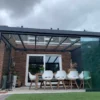 Nowoczesne patio ma szklany dach i ściany, długi stół jadalny z białymi i wiklinowymi krzesłami i łączy się z ceglanym domem z przesuwanymi drzwiami. Zadaszenie tarasowe Cube dodaje stylu, a na zewnątrz widać zieloną trawę i bluszcz.