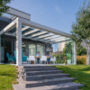 Stylowe przeszklone patio z białą pergolą Zadaszenie tarasowe Cube dołączone do domu, gdzie ludzie spożywają posiłki. Przestrzeń otoczona jest zieloną trawą, drzewami i roślinami krajobrazowymi pod czystym, błękitnym niebem.