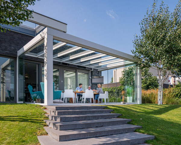 Stylowe przeszklone patio z białą pergolą Zadaszenie tarasowe Cube dołączone do domu, gdzie ludzie spożywają posiłki. Przestrzeń otoczona jest zieloną trawą, drzewami i roślinami krajobrazowymi pod czystym, błękitnym niebem.