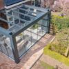 Zadaszenie tarasowe Cube to stylowa pergola z przeszkloną obudową, tworząca przedłużenie patio z meblami jadalnymi, przymocowana do murowanego domu i wychodząca na zagospodarowany ogród z drzewami, krzewami i wybrukowanym chodnikiem.