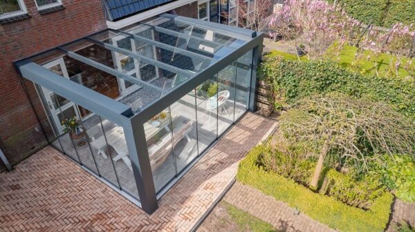 Zadaszenie tarasowe Cube to stylowa pergola z przeszkloną obudową, tworząca przedłużenie patio z meblami jadalnymi, przymocowana do murowanego domu i wychodząca na zagospodarowany ogród z drzewami, krzewami i wybrukowanym chodnikiem.