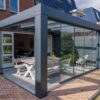 Zadaszenie tarasowe Cube, elegancka szklana pergola, przykrywa biały stół piknikowy i krzesła na wybrukowanym patio ceglanego domu, łącząc nowoczesny styl z bujnym widokiem na ogród.