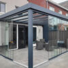 Zadaszenie tarasowe Cube to nowoczesna szklana obudowa patio przymocowana do domu, charakteryzująca się elegancką konstrukcją pergoli z ciemną metalową ramą i przesuwanymi szklanymi drzwiami, oferująca zachęcającą przestrzeń do relaksu.