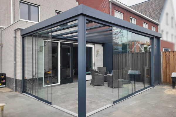 Zadaszenie tarasowe Cube to nowoczesna szklana obudowa patio przymocowana do domu, charakteryzująca się elegancką konstrukcją pergoli z ciemną metalową ramą i przesuwanymi szklanymi drzwiami, oferująca zachęcającą przestrzeń do relaksu.