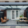 Zadaszenie tarasowe Cube prezentuje eleganckie, przeszklone patio z nowoczesną pergolą, opalonymi fotelami, drewnianą ławką i stołem z roślinami, poduszkami, przesuwanymi drzwiami z białymi żaluzjami i drewnianym ogrodzeniem graniczącym z przestrzenią.